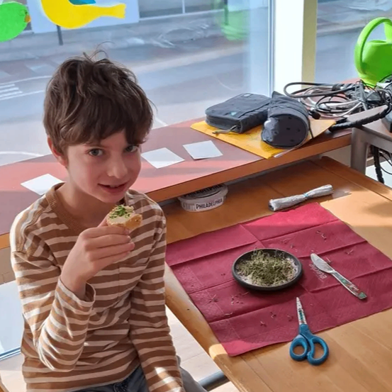 Ein junger Junge sitzt an einem Tisch und isst ein Sandwich, mit einer roten Tischdecke und einer Schere in der Nähe. Hinter ihm befindet sich ein Fenster mit farbigen Vogelausschnitten.