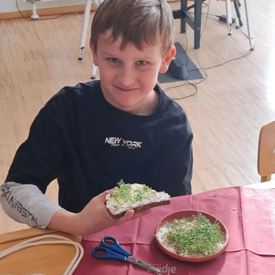 Ein Junge mit einem schwarzen T-Shirt sitzt an einem Tisch und hält ein Sandwich mit grünen Sprossen. Der Tisch hat ein rotes Tuch und einen Teller mit weiteren Sprossen. Hinter ihm steht eine Gitarre auf einem Ständer.
