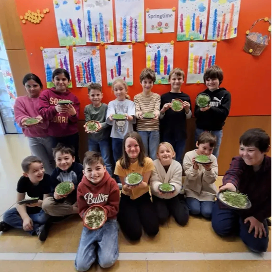 Eine Gruppe von Kindern posiert für ein Foto, während sie Teller mit grünem Gras halten. Sie tragen alle verschiedenfarbige Hemden. An der Wand hinter ihnen sind ein paar Papiere befestigt, und rechts steht ein Korb.
