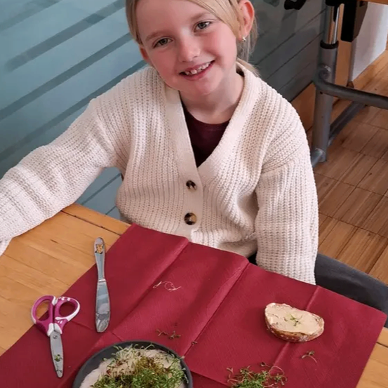 Ein junges Mädchen mit blonden Haaren sitzt an einem holzfarbenen Tisch mit einem roten Tuch und lächelt. Auf dem Tisch befinden sich ein Gericht mit Sprossen, ein Stück Brot und eine Schere.