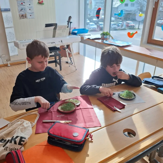 Zwei Jungen sitzen an einem Tisch mit Essen. Einer hält ein Messer und schaut auf sein Essen. Der andere Junge isst und schaut auf etwas. Ein Klavier, eine Pflanze und Taschen stehen auf dem Tisch. Ein Auto ist draußen geparkt.