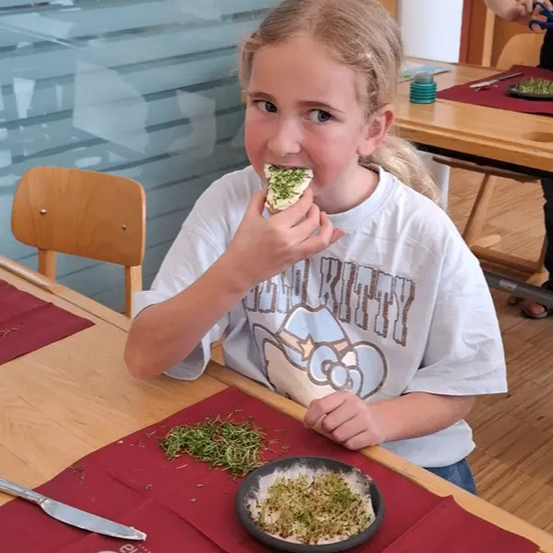 Ein junges Mädchen isst einen grün bedeckten Lebensmittelartikel an einem Tisch mit rotem Tuch, Scheren und Messer.