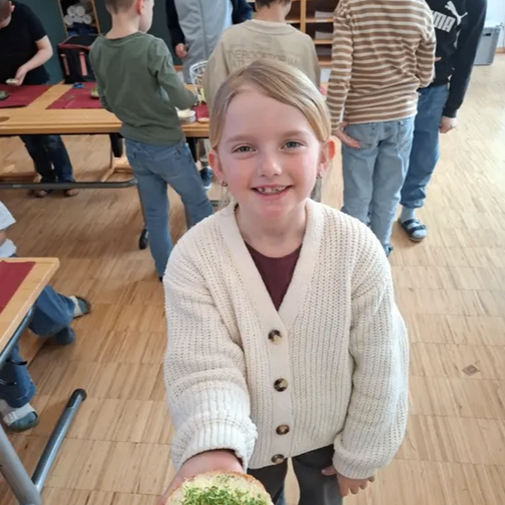 Ein junges Mädchen in einem weißen Strickjacke hält ein Sandwich mit grünen Toppings. Hinter ihr stehen andere Kinder und arbeiten an einem Tisch an Aufgaben.