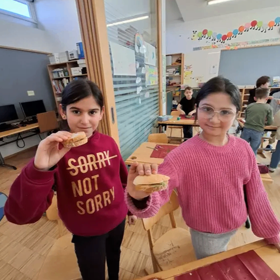 Zwei Mädchen in einem Klassenzimmer, eines in einem weinroten Pullover, das andere in einem rosa Pullover, beide halten Sandwiches, mit einem Tisch und Stühlen vor ihnen.