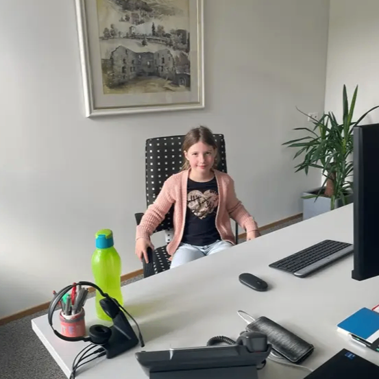 Ein junges Mädchen sitzt in einem Büro an einem Schreibtisch, umgeben von verschiedenen Gegenständen wie Tastatur, Maus und einer grünen Wasserflasche. Hinter ihr hängt ein gerahmtes Bild an der Wand.