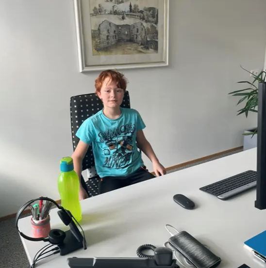 Ein Junge sitzt an einem Schreibtisch im Büro und schaut in die Kamera. Er trägt ein blaues T-Shirt. Auf dem Schreibtisch stehen eine grüne Wasserflasche, eine schwarze Maus, eine Tastatur und ein Cisco-Gerät.