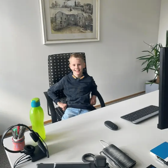 Ein junger Junge sitzt in einem Bürostuhl und lächelt. Der Schreibtisch hat einen Computer, eine Tastatur und verschiedene Gegenstände wie einen Stifthalter und eine Wasserflasche. Ein gerahmtes Bild hängt an der Wand.