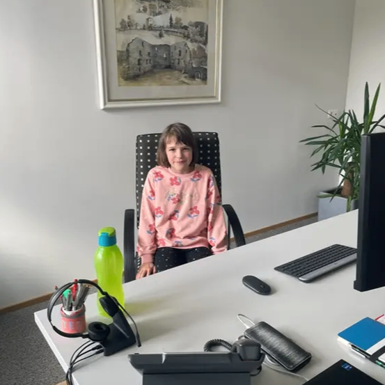 Ein Mädchen in einem rosa Shirt sitzt an einem Schreibtisch mit Computermonitor, Tastatur und Maus. Auf dem Schreibtisch stehen eine grüne Flasche und ein Becher mit Stiften. Ein gerahmtes Bild hängt an der Wand.