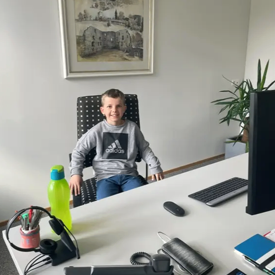 Ein junger Junge sitzt an einem Schreibtisch mit Computer, Tastatur und Maus, trägt ein Adidas Sweatshirt und lächelt. Der Schreibtisch ist mit einem Monitor, Kopfhörern, einer Wasserflasche und einem Topfpflanzen gefüllt.