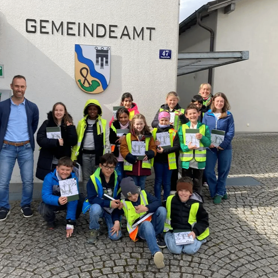 Eine Gruppe von Kindern und Erwachsenen posiert für ein Foto vor einem Gebäude. Sie halten Bücher und tragen Warnwesten. Das Gebäude hat ein Schild mit der Aufschrift 'GEMEINDEAMT'.