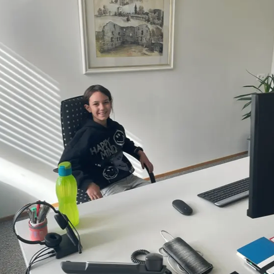 Ein Mädchen sitzt an einem Schreibtisch mit Computermonitor, Tastatur und einer Flasche. Ein gerahmtes Bild hängt an der Wand hinter ihr.