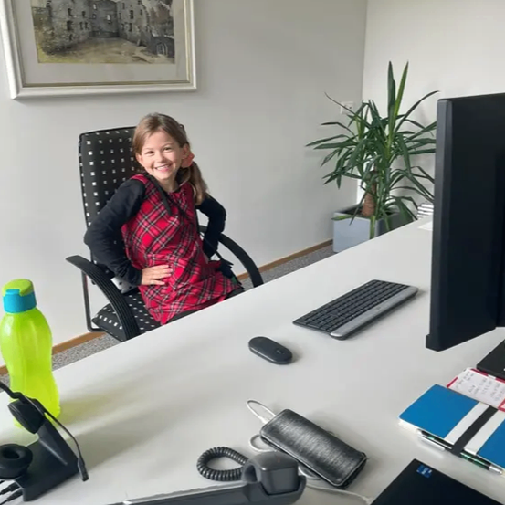 Ein junges Mädchen sitzt in einem Bürostuhl. Sie lächelt und posiert für ein Foto. Der Büroschreibtisch hat einen Monitor, eine Tastatur und andere Büroartikel.