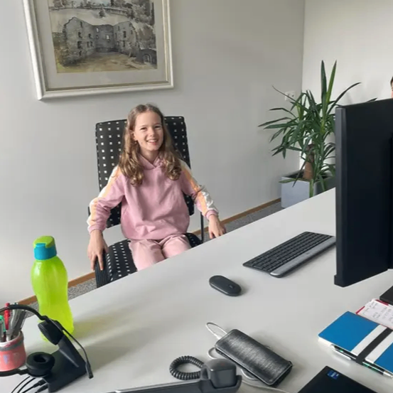 Ein junges Mädchen sitzt an einem Schreibtisch mit Computer, Tastatur und Maus. Sie lächelt und trägt eine rosa Jacke. Der Schreibtisch ist mit einem Telefon, einem Stifthalter und Büchern übersät. Ein Topf mit Pflanzen steht hinter ihr.