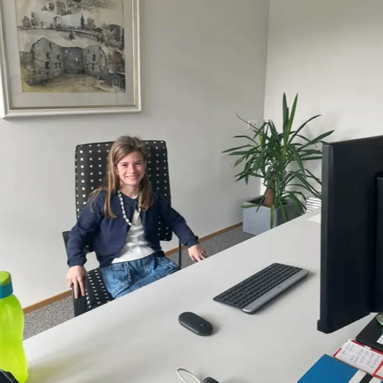 Eine junge Frau sitzt in einem Büro an einem Schreibtisch und lächelt. Der Schreibtisch hat einen Computer, eine Tastatur, eine Maus und andere Büroartikel. Ein Topf und ein gerahmtes Bild hängen an der Wand.