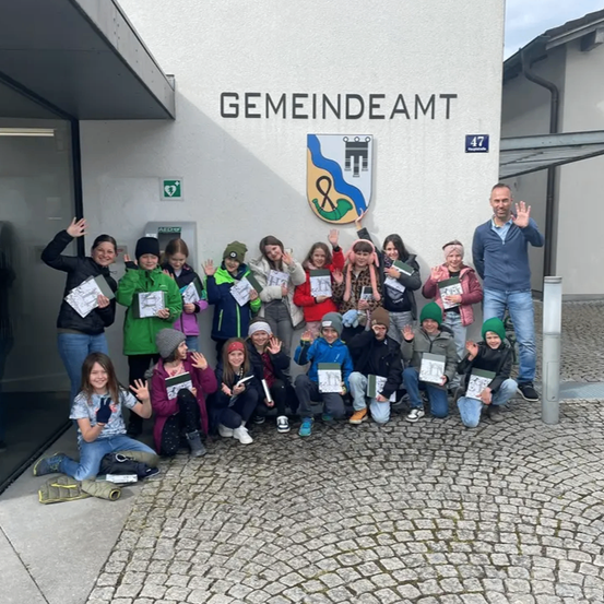 Eine Gruppe von Kindern und einem Mann posiert für ein Foto vor dem Gemeindeamt-Gebäude. Jedes Kind hält ein Zertifikat und sie alle lächeln.