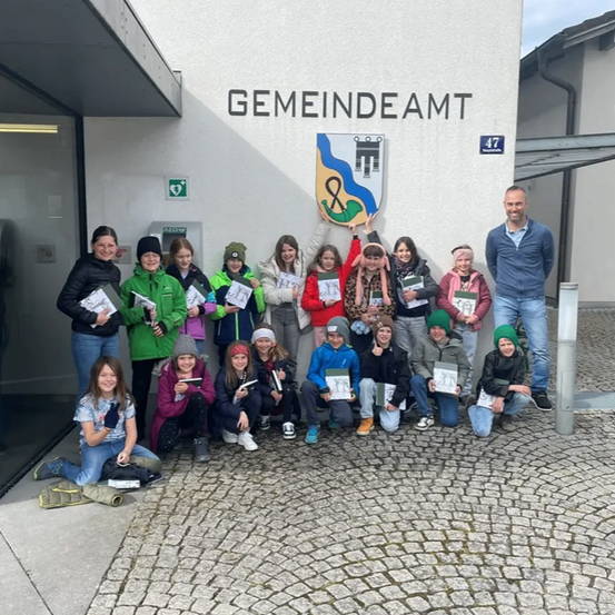Eine Gruppe von Kindern und Erwachsenen posiert für ein Foto vor einem Gebäude mit der Aufschrift 'GEMEINDEAMT' und hält Urkunden in den Händen. Ein Mann steht hinter ihnen.