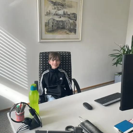 Ein junger Junge sitzt an einem Schreibtisch mit Computermonitor, Tastatur und Maus. Er trägt eine schwarze Jacke. Eine Pflanze, eine Wasserflasche und Kopfhörer stehen auf dem Schreibtisch.