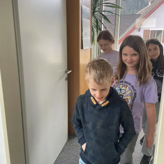 Vier Kinder stehen in einem Flur. Der Junge vorne hat die Hände in den Taschen und schaut zur Seite. Das Mädchen neben ihm lächelt und schaut nach vorne. Dahinter stehen ein weiteres Mädchen und ein Junge.