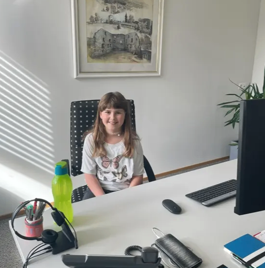 Ein junges Mädchen sitzt lächelnd und posiert für ein Foto an einem Schreibtisch im Büro. Auf dem Schreibtisch befinden sich ein Monitor, eine Tastatur, eine Maus und verschiedene Gegenstände wie eine Flasche, ein Becher mit Stiften und ein Notizbuch. Ein gerahmtes Bild hängt an der Wand hinter ihr.