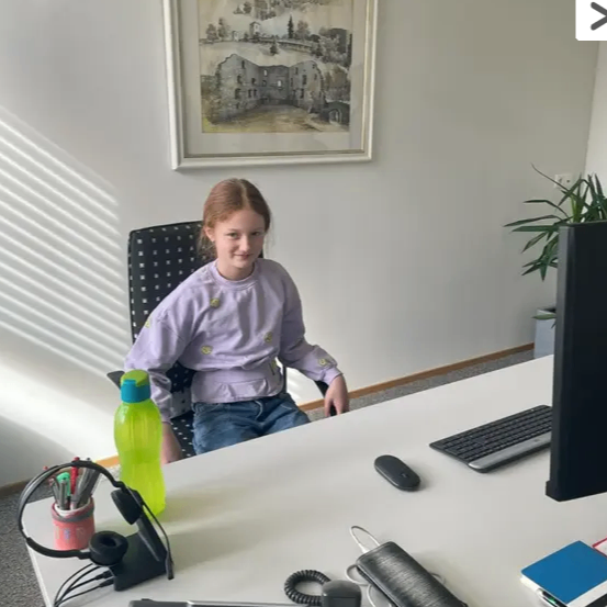 Ein Mädchen in einem lila Shirt sitzt an einem Schreibtisch mit einem Computer und einer Flasche. Ein Bild hängt an der Wand.