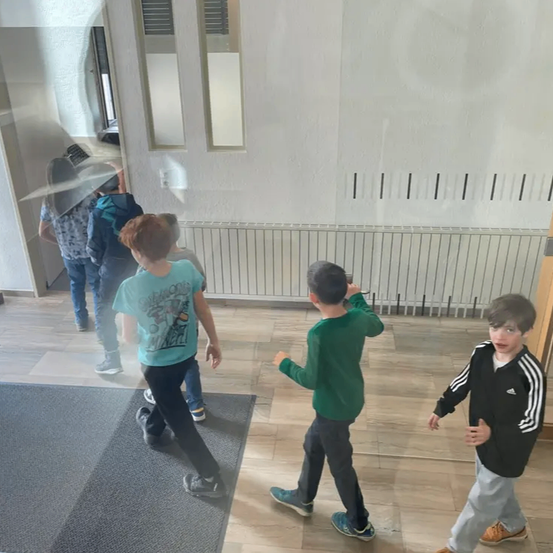 Eine Gruppe von Kindern geht in einem Raum mit Holzboden. Ein Kind hält einen Regenschirm. An der Wand steht ein Heizkörper.