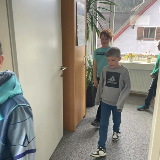 Drei Kinder gehen in einem Flur. Ein Junge in einem Adidas-Sweatshirt schaut nach unten. Hinter ihm ist ein Junge in einem grünen T-Shirt. Es gibt eine Topfpflanze und ein Fenster mit Fensterläden.