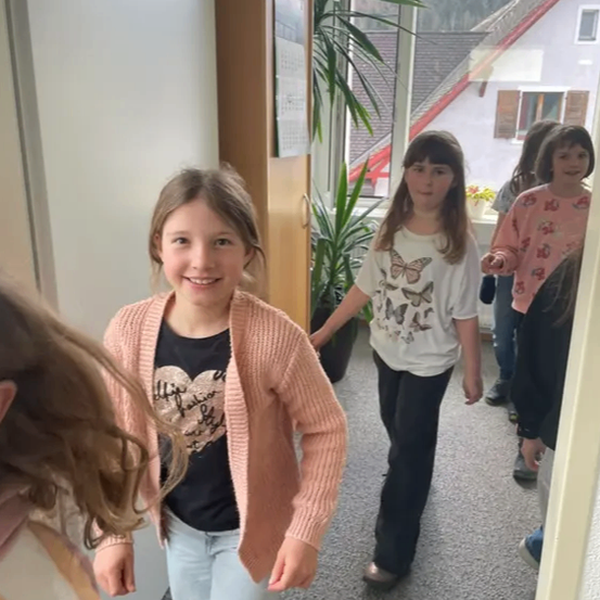 Mehrere junge Mädchen gehen durch einen Flur, wobei ein Mädchen in einem rosa Cardigan lächelt. Ein Topf mit Pflanzen und ein Haus mit rotem Rand sind im Hintergrund zu sehen.