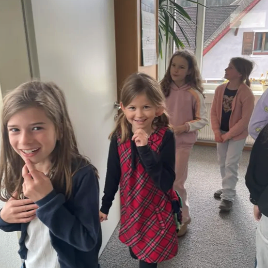 Fünf Mädchen sind drinnen, wahrscheinlich in einem Flur. Ein Mädchen trägt ein kariertes Kleid und ein anderes eine Jeansjacke. Sie scheinen für ein Foto zu posieren.