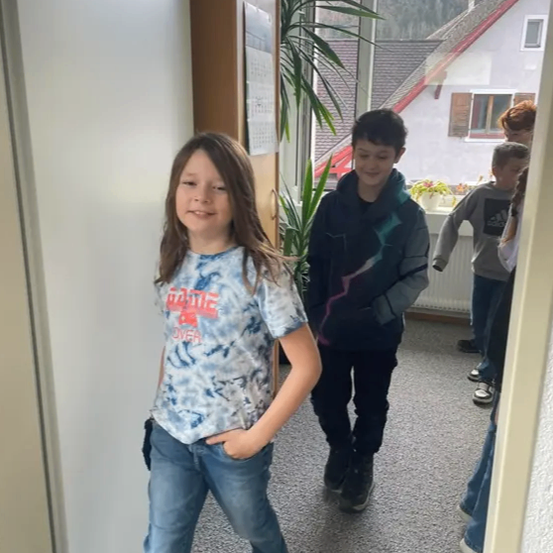 Drei Kinder gehen durch einen Flur mit einem Haus im Hintergrund. Ein Mädchen vorne trägt ein blau-weißes Tie-Dye-Shirt, während der Junge hinter ihr eine schwarz-grüne Jacke trägt.