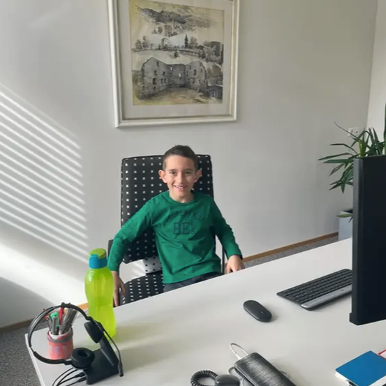 Ein Junge in einem grünen Shirt sitzt lächelnd an einem Schreibtisch, mit Tastatur, Maus und verschiedenen Büroartikeln auf dem Schreibtisch. Ein Bilderrahmen und eine Pflanze im Hintergrund.