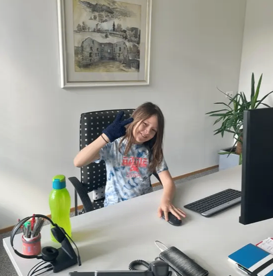 Ein junges Mädchen sitzt am Schreibtisch und lächelt. Hinter ihr ziert ein gerahmtes Bild die Wand. Der Schreibtisch ist mit einer Tastatur, einer Maus und einer grünen Wasserflasche übersät.