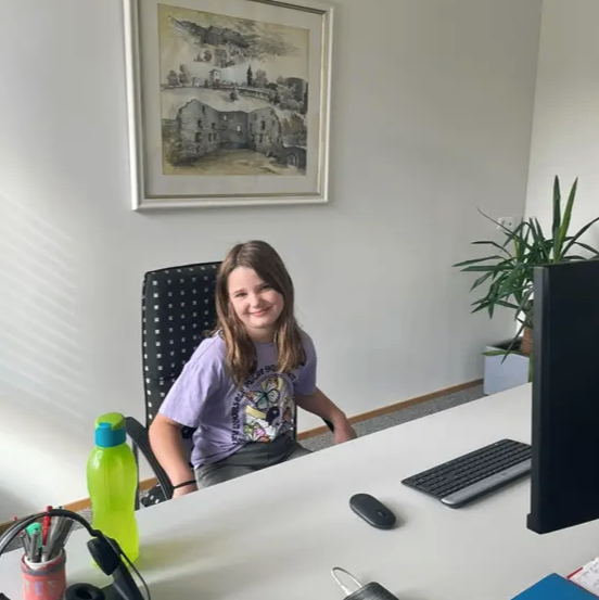 Ein Mädchen sitzt an einem Schreibtisch mit Computermonitor, Tastatur und einer Topfpflanze. Sie trägt ein lila T-Shirt. Auf dem Schreibtisch befinden sich eine grüne Flasche, Kopfhörer und ein Notizbuch.