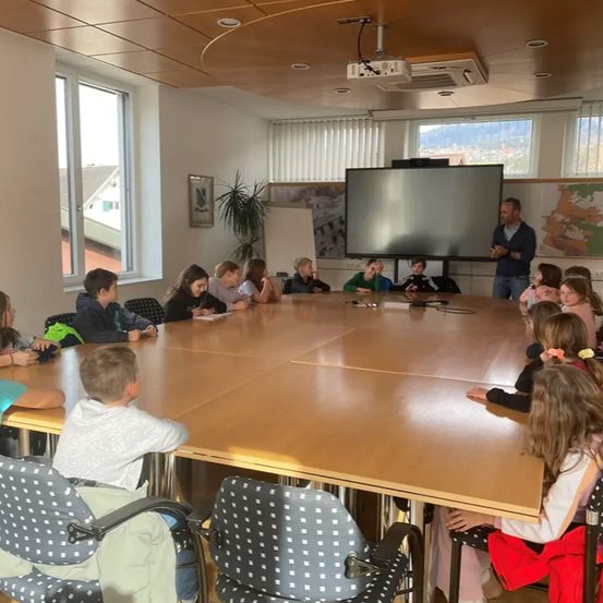 Ein Klassenzimmer mit Schülern und einem Lehrer, einer Projektionsleinwand und einer Tafel ist zu sehen. Ein Mann steht und spricht zu den Schülern, die aufmerksam sitzen. Der Raum ist gut beleuchtet mit Fenstern und einer Deckenventilation.