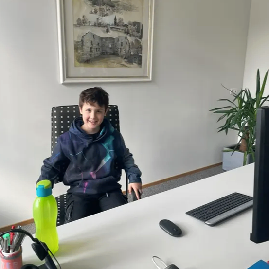 Ein junger Junge sitzt an einem Schreibtisch in einem Büro. Er lächelt und ist von Büromaterial umgeben. Ein gerahmtes Bild hängt an der Wand hinter ihm.