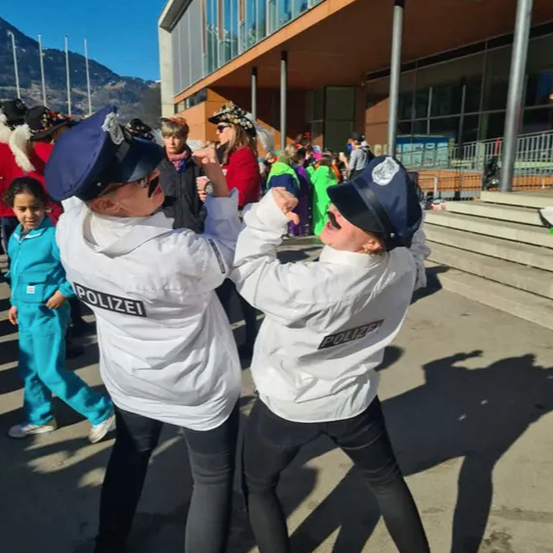 Zwei Frauen, die als Polizistinnen verkleidet sind, tanzen vor einem Gebäude, während im Hintergrund Menschen in verschiedenen Kostümen stehen.