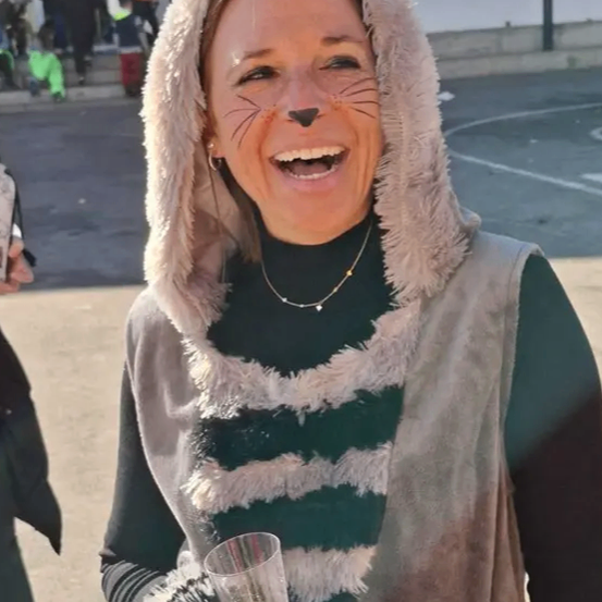 Eine Frau mit Katzen-Gesichtsbemalung steht draußen, lächelt, hält ein Weinglas und hat ein Gebäude und Menschen im Hintergrund.