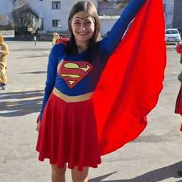 Eine Frau in einem Superman-Kostüm steht auf einem Parkplatz und hält ihren Umhang. Dahinter befindet sich ein Gebäude mit Bäumen und einem geparkten Auto vor der Fassade.
