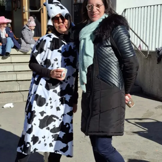 Zwei Frauen stehen zusammen, eine in einem Kuhkostüm und die andere in einem schwarzen Mantel. Sie lächeln und posieren für ein Foto.