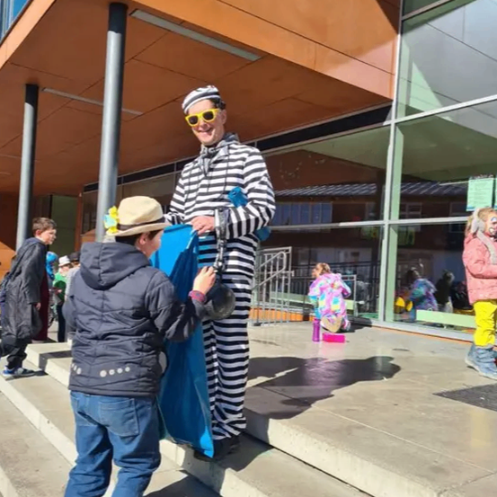 Ein Mann, der als Clown gekleidet ist, steht auf den Stufen eines Gebäudes. Er hält eine blaue Tasche und lächelt. Ein Kind steht neben ihm und hält die Tasche. Einige Leute stehen hinter ihnen auf den Stufen. Das Gebäude hat Glasfenster.