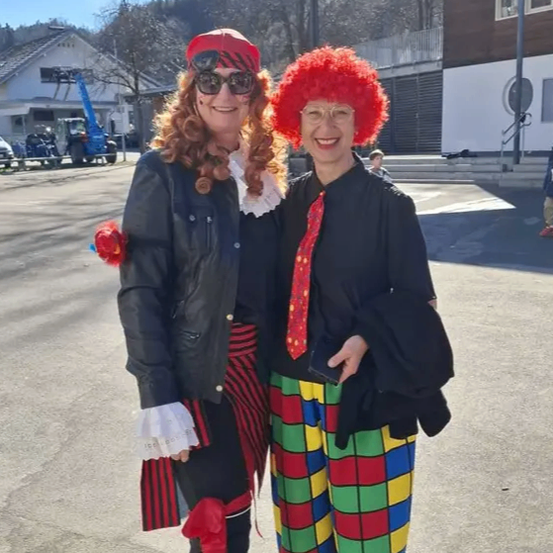 Zwei Frauen in Kostümen, eine in einem Piratenoutfit und die andere in einem Clownkostüm, stehen draußen auf einer Straße. Hinter ihnen befindet sich ein Gebäude, geparkte Fahrzeuge und eine Person, die geht.