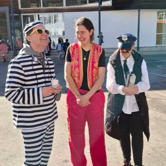 Drei Personen in Kostümen stehen vor einem Gebäude. Der Mann auf der linken Seite trägt ein gestreiftes Hemd und Sonnenbrille. Die Frau in der Mitte trägt eine Weste und rote Hosen. Der Mann auf der rechten Seite trägt einen Polizeihut und eine Maske.