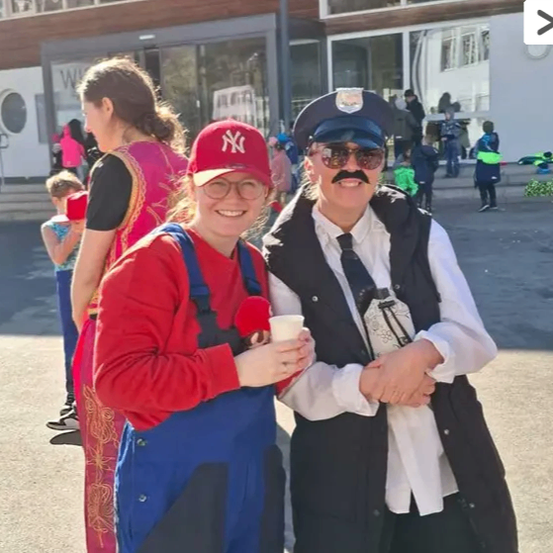 Zwei Menschen in Kostümen stehen draußen. Eine Frau in einer Mario-Verkleidung und ein Mann in einem Polizeiuniform. Sie halten beide Gegenstände und lächeln. Hinter ihnen sind andere Menschen und ein Gebäude.