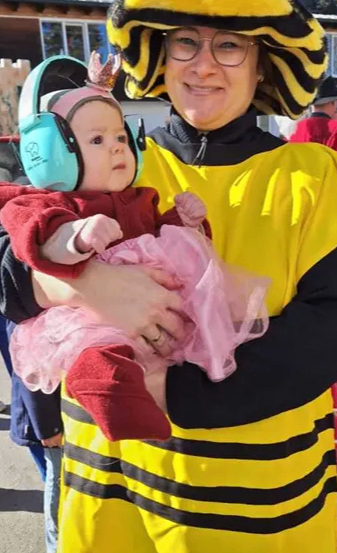 Eine Frau, die als Biene verkleidet ist, hält ein Baby, das einen rosa Tüllrock und eine Krone trägt und Geräuschunterdrückungs-Kopfhörer trägt. Im Hintergrund ist eine verschwommene Ansicht eines Gebäudes zu sehen.