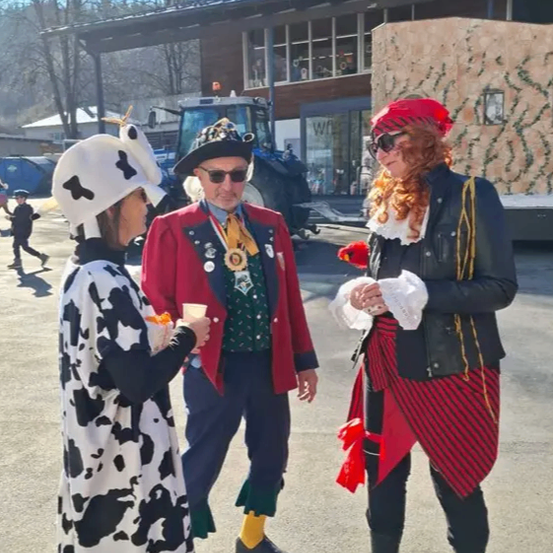 Drei Menschen in Kostümen, eine Kuh, ein Mann und ein Pirat, stehen draußen. Die Kuh hält einen Becher. Ein Traktor ist hinter ihnen geparkt.