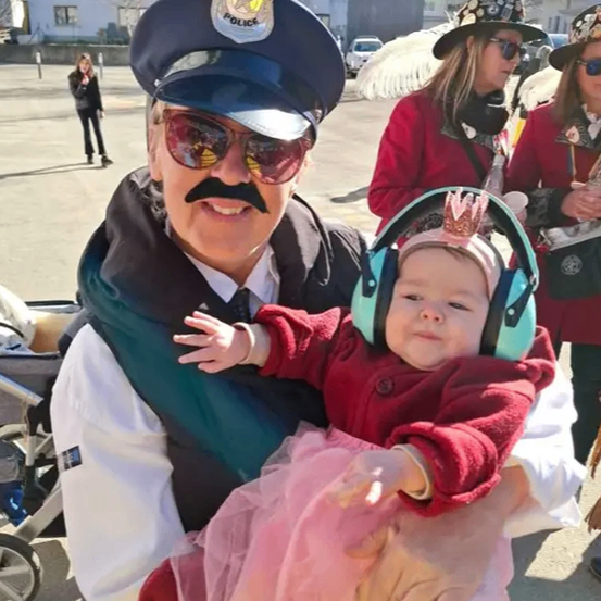 Ein Mann in einer Polizeiuniform hält ein Babymädchen in seinen Armen. Sie stehen im Freien. Hinter ihnen steht eine Frau mit einem Kinderwagen. Im Hintergrund befinden sich Gebäude und Autos.
