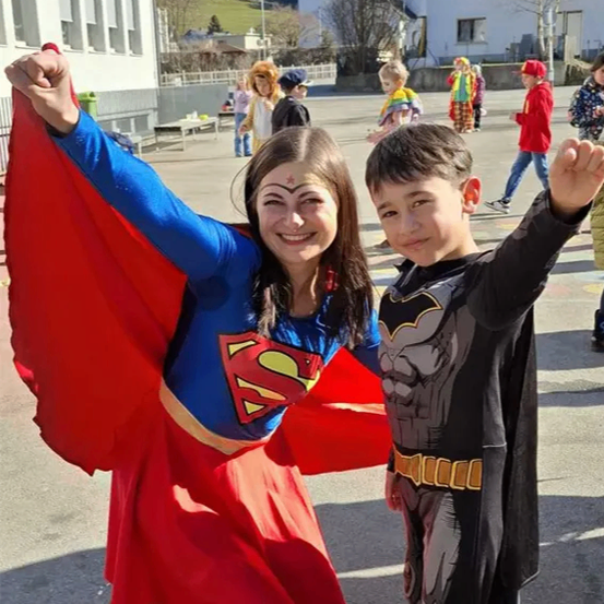 Ein junges Mädchen in einem Superman-Kostüm und ein Junge in einem Batman-Kostüm posieren für ein Foto im Freien. Hinter ihnen gehen Kinder in verschiedenen Kostümen herum. Im Hintergrund befinden sich Gebäude und Bäume.