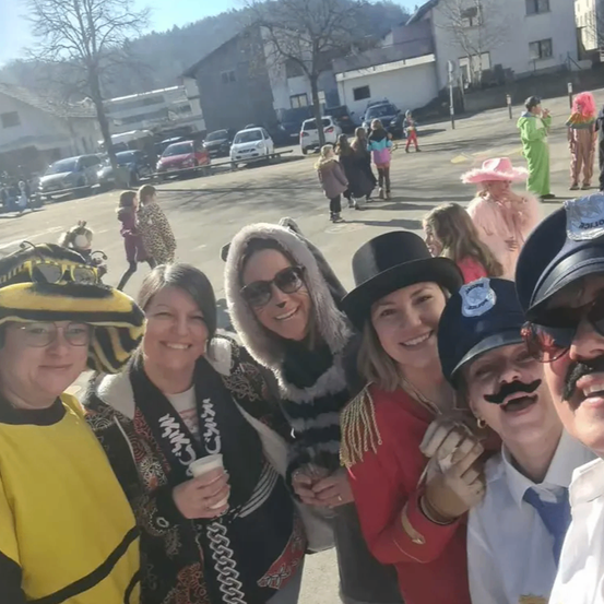 Eine Gruppe von Menschen in verschiedenen Kostümen, einschließlich Hüten und Brillen, steht zusammen an einem sonnigen Außenort. Dahinter gehen mehrere Menschen umher, einige ebenfalls in Kostümen. Im Hintergrund befinden sich Gebäude und geparkte Autos.