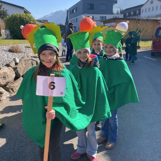 Kinder in grünen Kostümen mit Nummern auf den Hüten laufen in einer Parade. Der Hintergrund zeigt Häuser und Berge.
