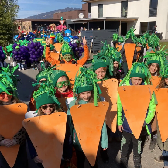 Eine Gruppe von Kindern, die als Karotten mit grünen Blättern auf dem Kopf verkleidet sind, stehen in einer Reihe draußen. Einige haben Ballons, und ein Gebäude steht dahinter.