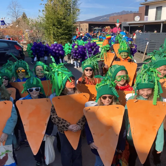 Eine Gruppe von Kindern in Karottenkostümen, die Karottenausschnitte halten, zieht in Kostümen mit grünen Blattkronen die Straße entlang.